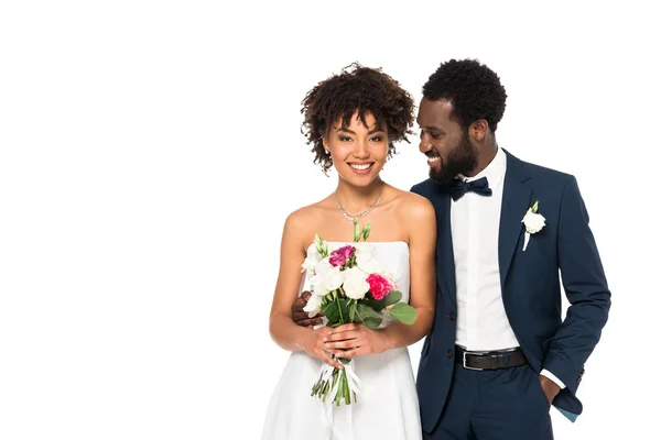 Feliz noiva afro-americana segurando buquê perto do noivo de pé com a mão no bolso isolado no branco — Fotografia de Stock