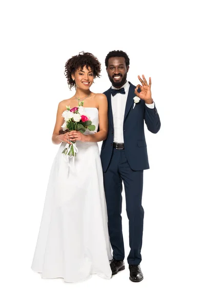 Noivo americano africano feliz mostrando sinal ok perto de noiva alegre segurando flores isoladas no branco — Fotografia de Stock