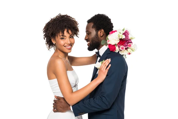 Sorridente sposa afro-americana che abbraccia lo sposo mentre tiene i fiori isolati sul bianco — Foto stock