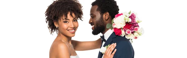 Tiro panorâmico de noiva americana africana feliz abraçando noivo enquanto segurando flores isoladas no branco — Fotografia de Stock