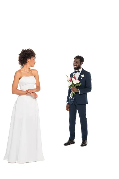 Noiva americana africana feliz de pé e olhando para noivo barbudo segurando flores isoladas no branco — Fotografia de Stock
