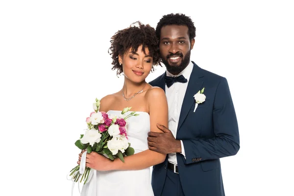 Feliz afroamericano novia sosteniendo flores cerca guapo novio mirando cámara aislado en blanco - foto de stock