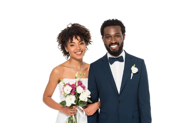 Joyeuse mariée afro-américaine tenant des fleurs près du marié en regardant la caméra isolée sur blanc — Photo de stock