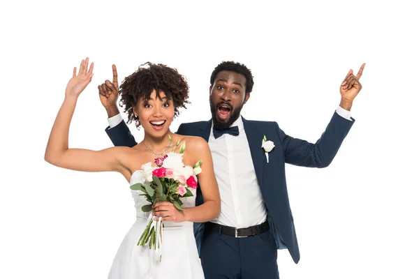 Happy african american bride standing with bridegroom and gesturing while holding flowers isolated on white — Stock Photo