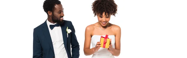 Plano panorámico de novio afroamericano feliz mirando alegre novia sosteniendo caja de regalo aislado en blanco - foto de stock