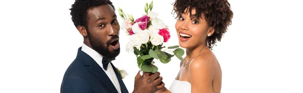 Plano panorámico de novio afroamericano sorprendido y novia feliz sosteniendo flores aisladas en blanco - foto de stock