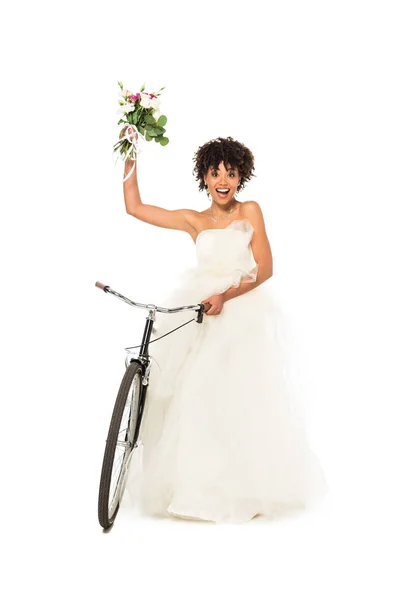 Joyeuse mariée afro-américaine en robe de mariée tenant des fleurs et vélo isolé sur blanc — Photo de stock