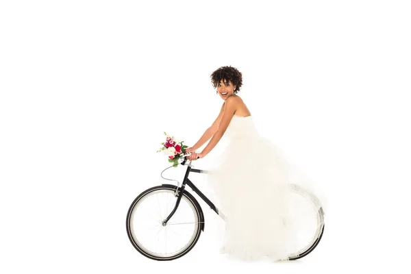 Alegre noiva afro-americana em vestido de noiva segurando flores enquanto andava de bicicleta isolada no branco — Fotografia de Stock
