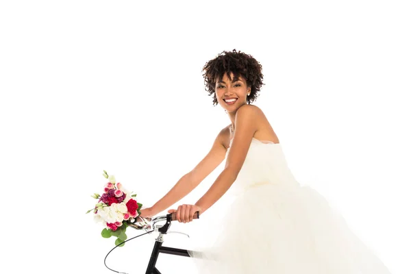 Glücklich afrikanisch-amerikanische Braut im Hochzeitskleid mit Blumen, während Fahrrad fahren isoliert auf weiß — Stockfoto