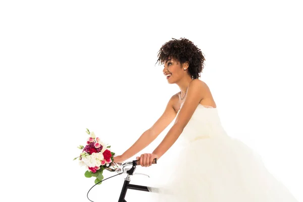 Low angle view of attractive african american bride in wedding dress holding flowers while riding bicycle isolated on white — Stock Photo