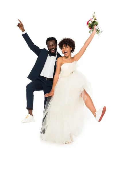 Joyeux marié afro-américain pointant du doigt près de mariée heureuse tenant des fleurs isolées sur blanc — Photo de stock