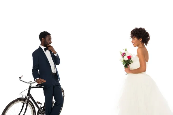 African american bridegroom standing near bicycle and looking at beautiful bride with flowers isolated on white — Stock Photo