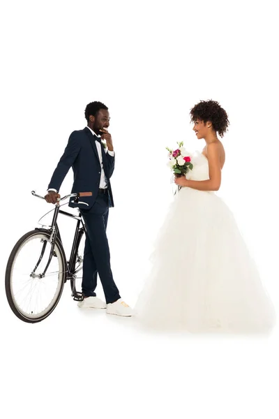 Novio afroamericano de pie cerca de la bicicleta y mirando a la novia alegre con flores aisladas en blanco - foto de stock