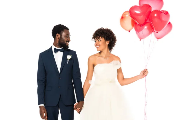 Allegro sposo afroamericano che si tiene per mano con attraente sposa vicino palloncini isolati su bianco — Foto stock