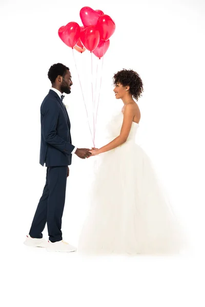 Vista lateral del alegre novio afroamericano cogido de la mano con atractiva novia cerca de globos aislados en blanco - foto de stock