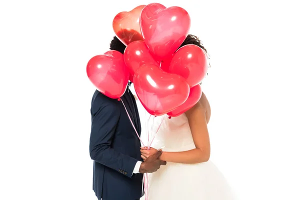 African american bridegroom and bride covering faces with pink heart-shaped balloons isolated on white — Stock Photo