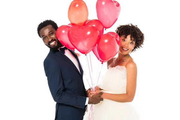 Allegro sposo africano americano che si tiene per mano con attraente sposa vicino palloncini rosa isolati su bianco — Foto stock