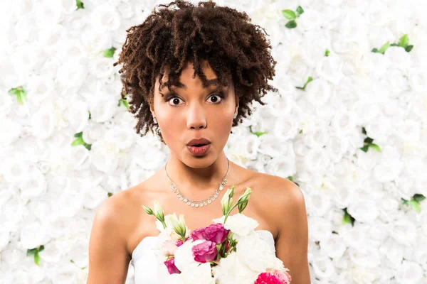 Sorprendido afroamericano novia mirando la cámara mientras sostiene flores - foto de stock