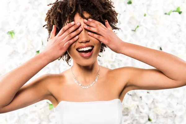 Fröhliche afrikanisch-amerikanische Braut bedeckt Augen mit Händen in der Nähe von Blumen — Stockfoto