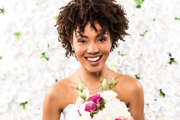 Mariée afro-américaine pocitive regardant la caméra tout en tenant des fleurs — Photo de stock