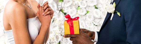 Panoramic shot of african american man giving present to bride near flowers — Stock Photo