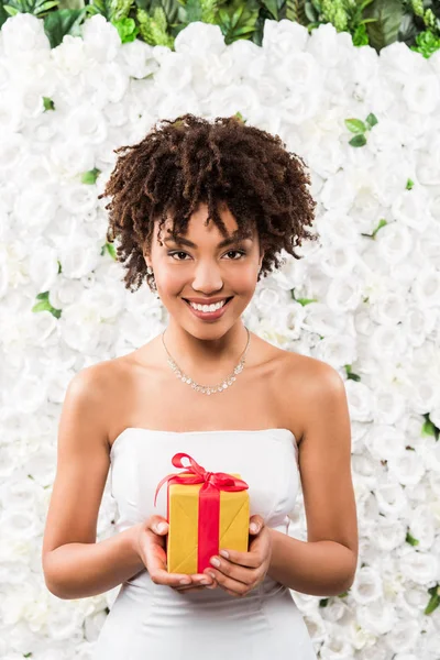 Fröhliche lockige afrikanisch-amerikanische Braut hält Geschenk in der Nähe von Blumen — Stockfoto
