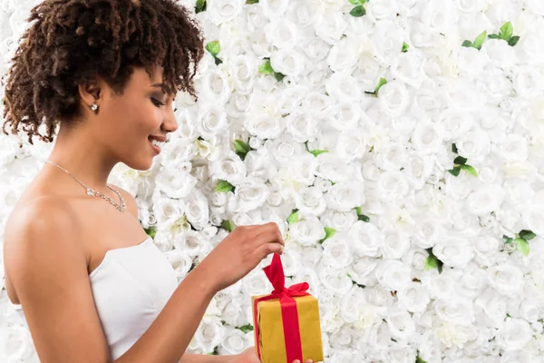 Side view of cheerful curly african american bride holding present near flowers — Stock Photo