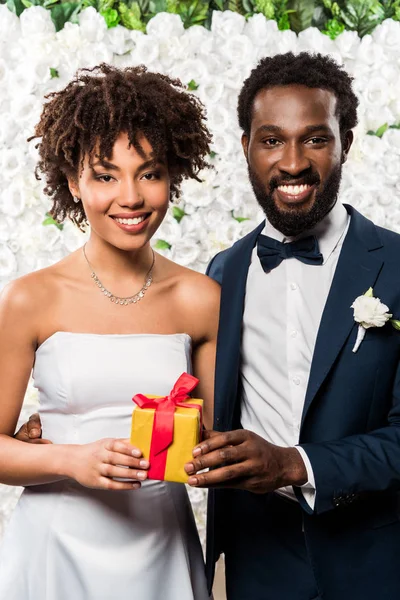 Heureuse mariée afro-américaine tenant présent près marié et fleurs — Photo de stock