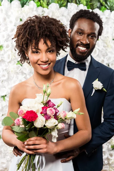 Cheerful african american bride holding bouquet with flowers near bridegroom — Stock Photo