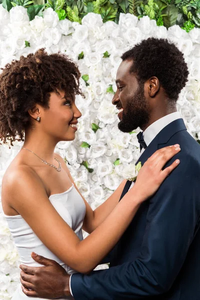 Feliz africano americano novia abrazando guapo barbudo novio cerca de flores - foto de stock