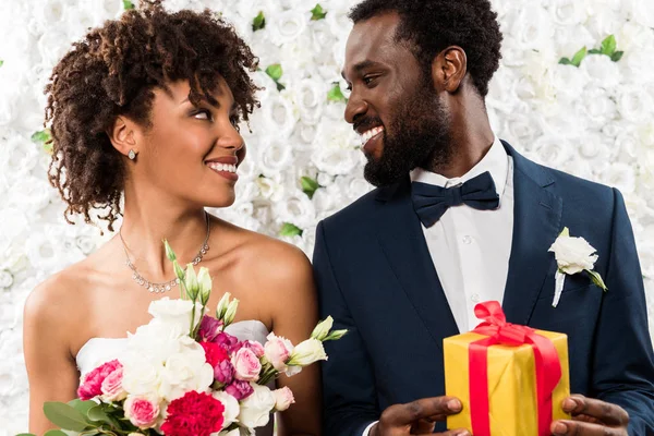 Cheerful african american bride holding fowers near happy bridegroom with present — Stock Photo