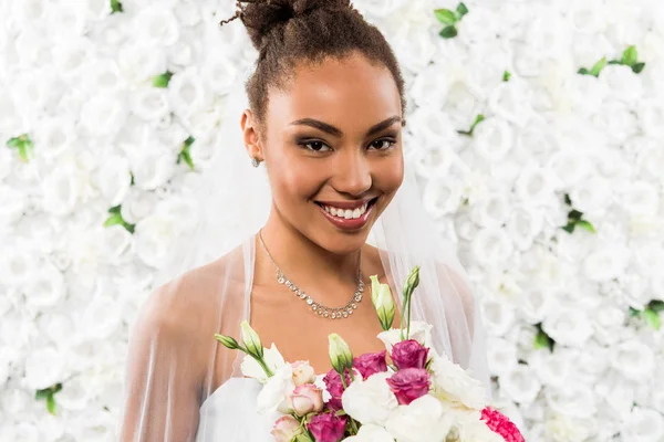 Cheerful african american bride in veil holding bouquet of flowers — Stock Photo