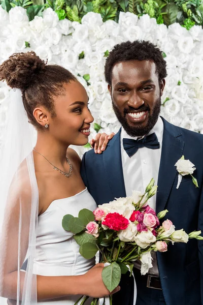 Noiva americana africana feliz em véu branco sorrindo perto do noivo e flores — Fotografia de Stock