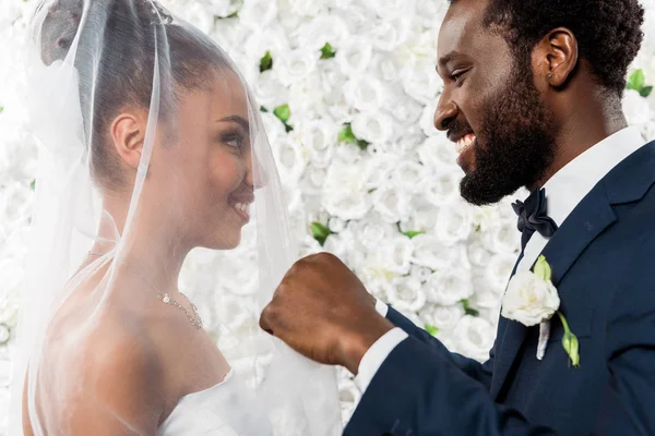 Felice sposo africano americano toccando velo bianco e sorridente vicino a sposa e fiori — Foto stock