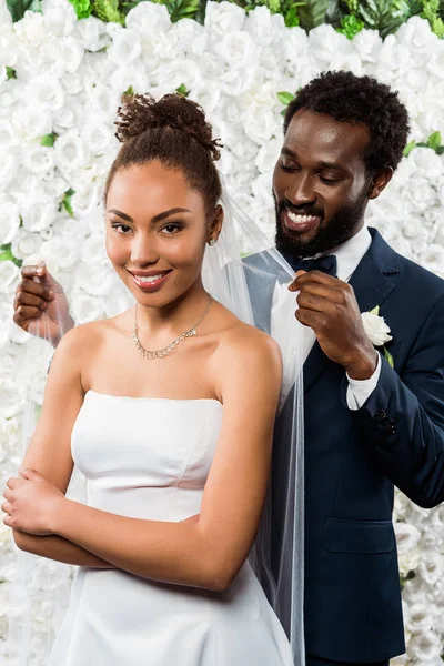 Fröhlich afrikanisch-amerikanischer Bräutigam berührt weißen Schleier und lächelt neben Braut und Blumen — Stockfoto