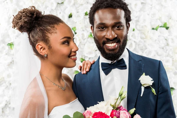 Счастливая африканская американская невеста смотрит на жениха, держащего цветы — стоковое фото