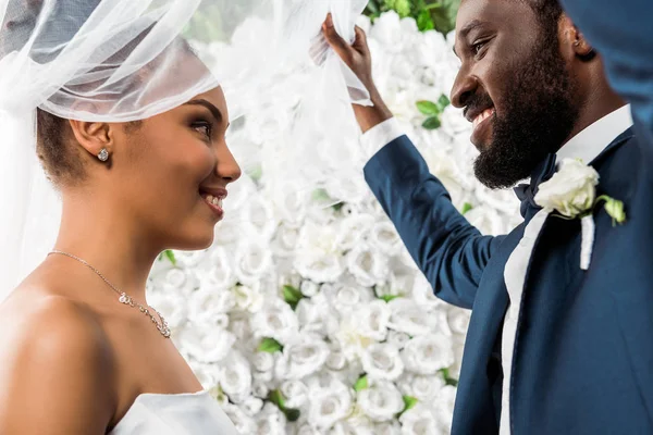 Vista de ángulo bajo del novio afroamericano feliz tocando el velo blanco y sonriendo cerca de novia y flores - foto de stock