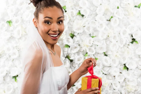 Überraschte afrikanisch-amerikanische Braut hält Geschenk in der Nähe von Blumen — Stockfoto