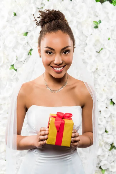 Cheerful african american bride in veil holding present near flowers — Stock Photo