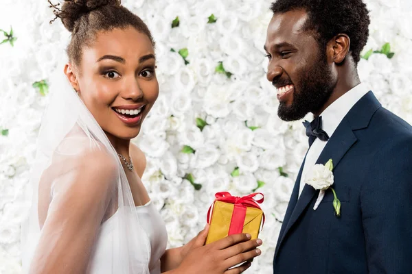 Noiva americana africana surpreso segurando presente perto do noivo feliz e flores — Fotografia de Stock