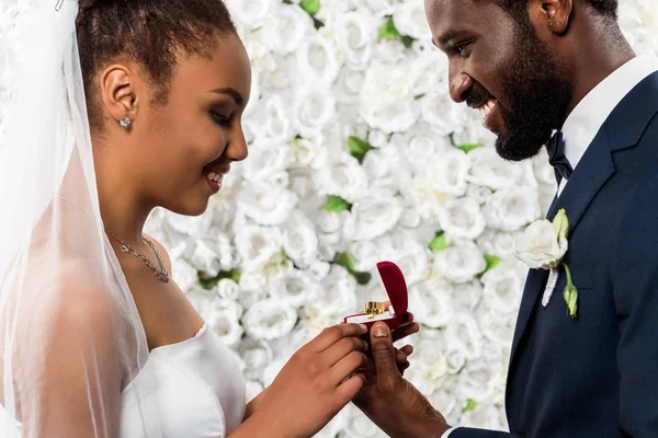 Cheerful african american bride looking at box with ring near happy bridegroom and flowers — Stock Photo
