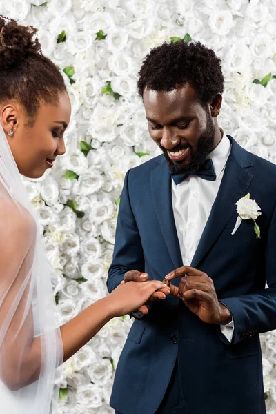 Alegre afroamericano hombre poner anillo de bodas en dedo de novia cerca de flores - foto de stock