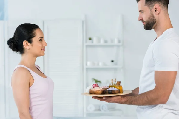 Guapo masajista sosteniendo bandeja con sal marina y aceite en botellas cerca de atractiva mujer - foto de stock