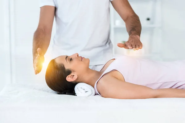 Vista recortada de curandero poner las manos por encima del cuerpo de la mujer atractiva con los ojos cerrados acostado en la mesa de masaje - foto de stock