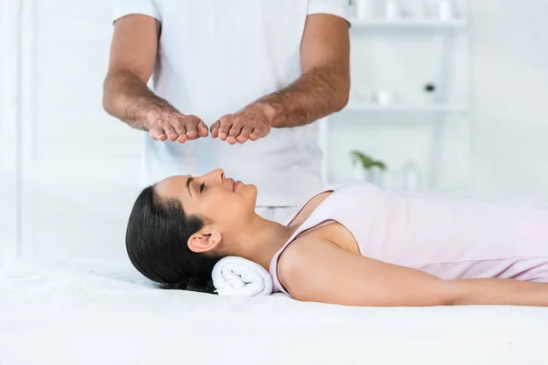 Vue recadrée du guérisseur mettre les mains au-dessus de la tête de la femme attrayante avec les yeux fermés couché sur la table de massage — Photo de stock