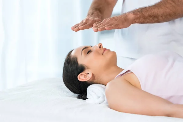 Cropped view of man putting hands above head of attractive woman with closed eyes lying on massage table — Stock Photo