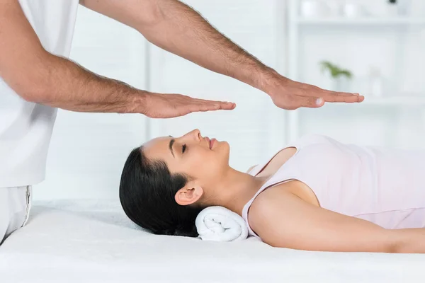 Vista recortada de curandero poner las manos por encima del cuerpo de la mujer con los ojos cerrados acostado en la mesa de masaje - foto de stock