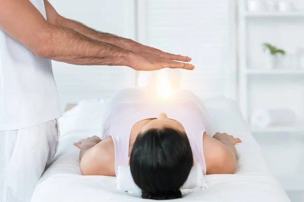 Vue recadrée du guérisseur mettant les mains au-dessus du corps de la femme brune allongée sur une table de massage — Photo de stock