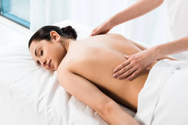 Vista cortada do massagista fazendo massagem à mulher morena deitada na mesa de massagem — Fotografia de Stock