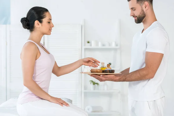 Atractiva morena mujer tomando botella con aceite mientras masajista celebración bandeja - foto de stock